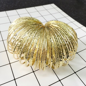 May include: A collection of gold-colored bangles arranged in a heart shape. The bangles have a textured surface and are stacked together. The background is a white surface with a black grid pattern.