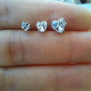 May include: Three silver-toned stud earrings with clear, heart-shaped cubic zirconia stones. The earrings are displayed in varying sizes, with the largest one on the right. The stones are faceted to catch the light, creating a sparkling effect.