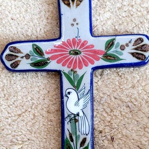May include: A white ceramic cross with blue trim and painted floral and bird designs. The cross features a pink flower with a yellow center, green leaves, and a white dove with brown accents.