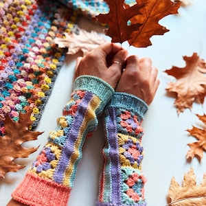Könnte beinhalten: Ein Paar bunte, gehäkelte Fingerhandschuhe. Die Handschuhe weisen eine Mischung aus Pastellfarben auf, darunter Rosa, Gelb, Blau und Lila. Die Manschetten sind einfarbig und der Hauptteil hat ein strukturiertes, gemustertes Design. Herbstblätter sind im Hintergrund.