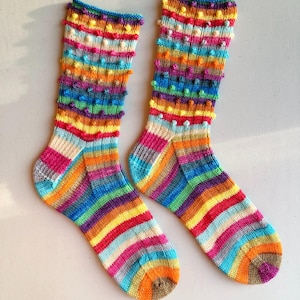 Könnte beinhalten: Ein Paar bunt handgestrickte Socken. Die Socken haben ein gestreiftes Muster in verschiedenen Farben, darunter Rosa, Orange, Gelb, Grün und Blau. Die Oberseite der Socken hat ein strukturiertes Noppenmuster. Die Socken sind aus weichem Garn gefertigt.