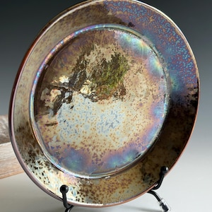 May include: A handmade ceramic plate with a unique iridescent glaze. The plate features a wide rim and a shallow center. The glaze has a mix of colors, including gold, brown, blue, and purple. The plate is displayed on a black metal stand.
