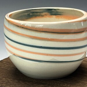 May include: A white ceramic bowl with a swirling pattern of blue and orange stripes. The bowl is sitting on a wooden surface.