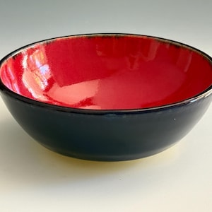 May include: A ceramic bowl with a glossy red interior and a dark blue exterior. The rim has a thin, dark line. The bowl is round and sits on a white surface. This bowl is suitable for serving food.