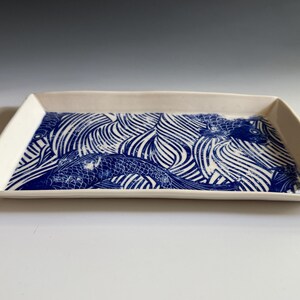 Pu&ograve; includere: Un vassoio rettangolare in ceramica con base bianca e un motivo grafico blu che raffigura pesci koi che nuotano tra linee d'acqua stilizzate. Il vassoio presenta bordi leggermente rialzati e una finitura liscia e lucida su tutta la superficie.