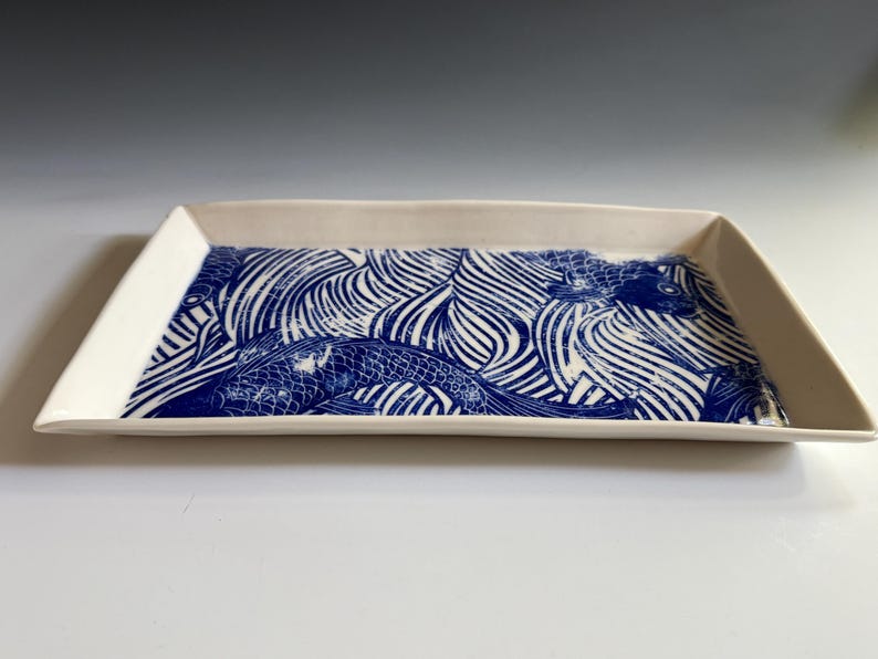 Pu&ograve; includere: Un vassoio rettangolare in ceramica con base bianca e un motivo grafico blu che raffigura pesci koi che nuotano tra linee d'acqua stilizzate. Il vassoio presenta bordi leggermente rialzati e una finitura liscia e lucida su tutta la superficie.
