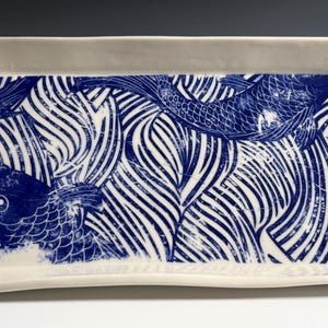 Pu&ograve; includere: Un vassoio rettangolare in ceramica con base bianca e una stampa grafica blu che raffigura pesci koi che nuotano tra motivi d'acqua stilizzati. Il vassoio presenta bordi rialzati e una finitura liscia e lucida su tutta la superficie.