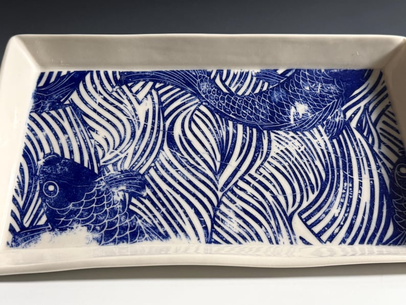 Pu&ograve; includere: Un vassoio rettangolare in ceramica con base bianca e una stampa grafica blu che raffigura pesci koi che nuotano tra motivi d'acqua stilizzati. Il vassoio presenta bordi rialzati e una finitura liscia e lucida su tutta la superficie.
