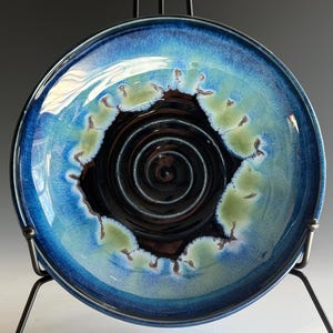 May include: A handmade ceramic plate with a glossy finish. The plate features a dark blue outer rim, transitioning to a lighter blue and green center, with a dark brown spiral design in the middle. The plate is displayed on a black metal stand.