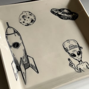 May include: A white ceramic square tray with black and white illustrations of a rocket, a moon, a UFO, and an alien wearing a baseball cap. The alien is pointing with one finger.