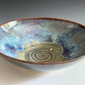 May include: A shallow ceramic bowl featuring a reactive glaze with blended shades of deep blue, purple, and earthy green. The interior base displays a distinct dark brown spiral pattern, while the outer rim is finished with a thin, warm brown edge.