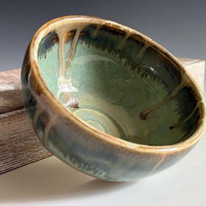 May include: A round ceramic bowl featuring a light sage green interior glaze with darker brown and tan drip patterns cascading from the rim. The exterior displays a similar blend of earthy brown and green tones with a smooth, glossy finish.