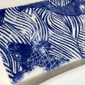 Pu&ograve; includere: Un vassoio rettangolare in ceramica con un motivo ad alto contrasto blu e bianco. Il design raffigura pesci koi stilizzati che nuotano tra correnti d'acqua lineari e fluide, resi con un'estetica a inchiostro usurato su una superficie bianca lucida.