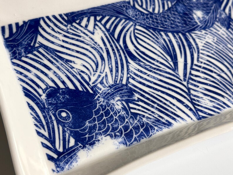 Pu&ograve; includere: Un vassoio rettangolare in ceramica con un motivo ad alto contrasto blu e bianco. Il design raffigura pesci koi stilizzati che nuotano tra correnti d'acqua lineari e fluide, resi con un'estetica a inchiostro usurato su una superficie bianca lucida.
