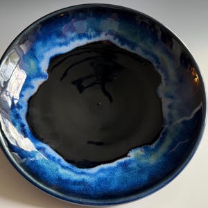 May include: A round ceramic bowl with a glossy finish. The bowl features a dark blue center that fades into a lighter blue and white rim. The outer edge is a darker blue.