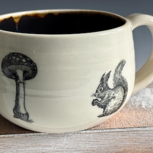 May include: Cream-colored ceramic mug with a dark brown interior and a curved handle. The mug features black and white illustrations of a mushroom and a squirrel. The mug is designed for hot beverages.