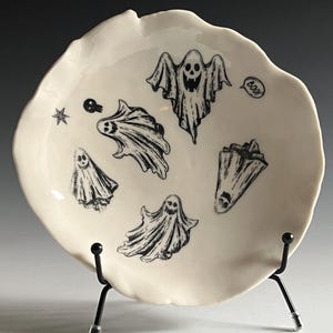 May include: White ceramic plate with a black and white ghost design. The plate has a scalloped edge and is on a black metal stand. The plate says "Boo!" in a speech bubble.