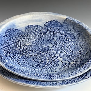 May include: Two ceramic plates with a blue and white doily pattern. The plates are stacked, with the top plate slightly larger than the bottom one. The doily design is intricate and covers the entire surface of the plates.