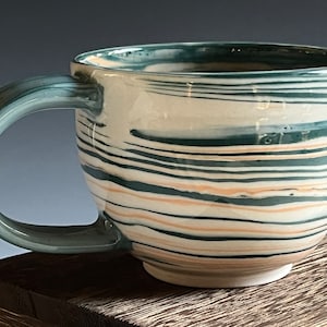May include: A ceramic mug with a white base and a swirling pattern of green and peach stripes. The mug has a rounded shape and a handle.