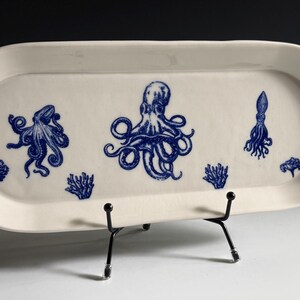 May include: An oval-shaped white ceramic serving platter featuring blue ink-style illustrations of two octopuses, a squid, and two coral formations. The platter is displayed on a black metal wire stand, showcasing a smooth, glossy finish.