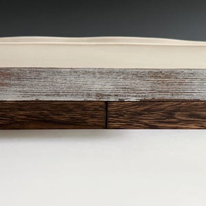 Pu&ograve; includere: Un vassoio rettangolare con una superficie superiore in ceramica liscia color bianco sporco, una sezione centrale in legno verniciato bianco anticato con venature visibili e una base composta da due blocchi di legno marrone scuro.