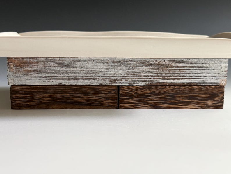 Pu&ograve; includere: Un vassoio rettangolare con una superficie superiore in ceramica liscia color bianco sporco, una sezione centrale in legno verniciato bianco anticato con venature visibili e una base composta da due blocchi di legno marrone scuro.