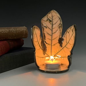 May include: A ceramic candle holder shaped like a leaf with a white glaze and black veins. A small tea light candle is burning inside the holder.