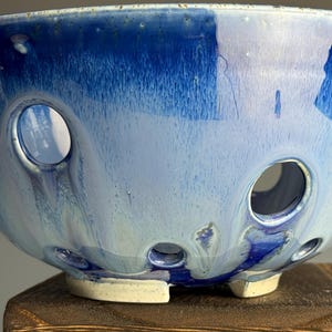 May include: A handcrafted ceramic bowl with a striking blue glaze. The bowl features a series of circular openings around its base and sides. The color transitions from a deep blue at the top to a lighter shade towards the center, creating a unique decorative piece.