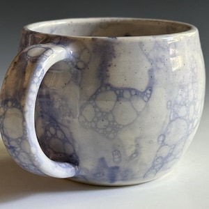 May include: A ceramic mug with a rounded body and a curved handle, featuring a white base glaze decorated with a light purple bubble pattern. The surface has a speckled texture and a glossy finish, showcasing a distinct circular bubble design.