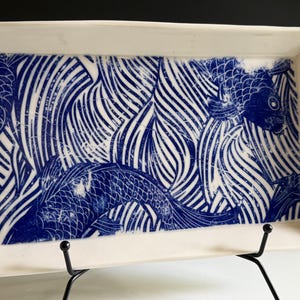 May include: A rectangular white ceramic tray featuring a deep blue graphic print of koi fish swimming amidst stylized flowing water patterns. The design covers the interior surface of the tray, which has raised edges and a smooth, glossy finish.