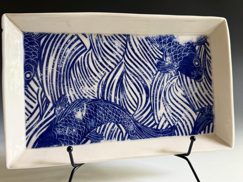 Pu&ograve; includere: Un vassoio rettangolare in ceramica bianca con una stampa grafica blu intenso raffigurante pesci koi che nuotano tra motivi d'acqua stilizzati. Il disegno copre la superficie interna del vassoio, dotato di bordi rialzati e finitura liscia.