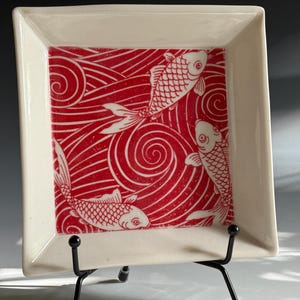 May include: Square ceramic dish with a white border and a red and white koi fish design. The center features a red background with white swirling lines and three white koi fish. The dish is displayed on a black metal stand.