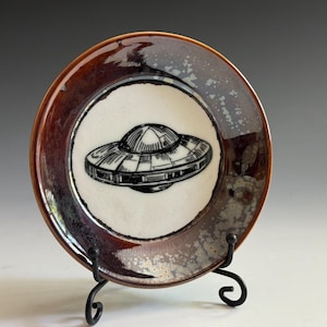 May include: A round ceramic plate featuring a black line drawing of a flying saucer in the center. The plate has a white circular interior surrounded by a wide, glossy rim with mottled brown, black, and metallic gray glaze effects.
