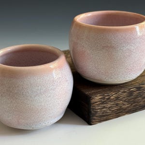 May include: Two small, round ceramic cups with a speckled pink and white glaze. The rims have a darker pink hue. One cup sits on a dark wooden block. The cups have a smooth, glossy finish.