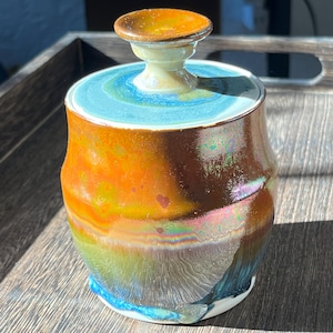 May include: A ceramic jar with a lid, featuring a colorful glaze with shades of orange, brown, green, and blue. The lid has a small, rounded knob on top.