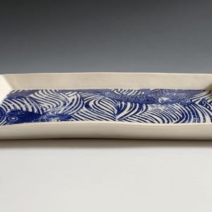 Pu&ograve; includere: Un vassoio rettangolare in ceramica con esterno color crema e base piatta. La superficie interna presenta un dettagliato motivo grafico blu e bianco che raffigura pesci stilizzati che nuotano tra linee curve e fluide su tutto il vassoio.