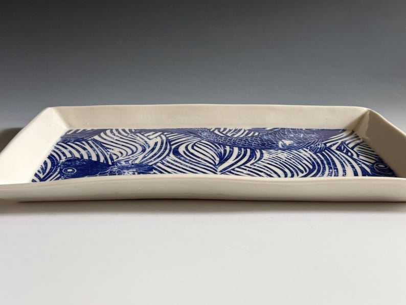 Pu&ograve; includere: Un vassoio rettangolare in ceramica con esterno color crema e base piatta. La superficie interna presenta un dettagliato motivo grafico blu e bianco che raffigura pesci stilizzati che nuotano tra linee curve e fluide su tutto il vassoio.