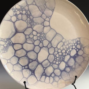 May include: A round ceramic plate with a cream-colored base featuring a blue bubble-like pattern that clusters across the surface. The design consists of various circular shapes of differing sizes, creating a dense, organic, and interconnected network.