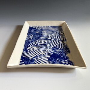 Pu&ograve; includere: Un vassoio rettangolare in ceramica con base color crema e una stampa grafica blu intenso. Il design presenta pesci koi stilizzati che nuotano tra linee ondulate e fluide che coprono la superficie interna del vassoio.