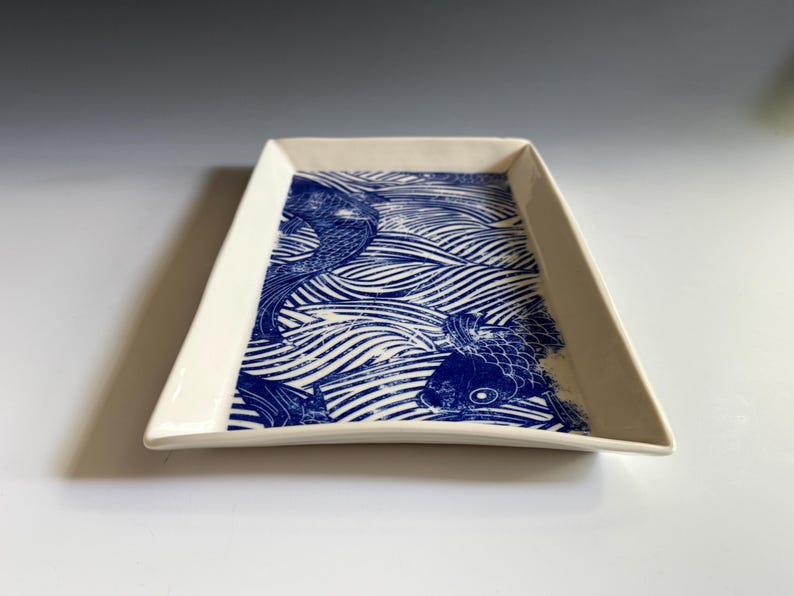 Pu&ograve; includere: Un vassoio rettangolare in ceramica con base color crema e una stampa grafica blu intenso. Il design presenta pesci koi stilizzati che nuotano tra linee ondulate e fluide che coprono la superficie interna del vassoio.