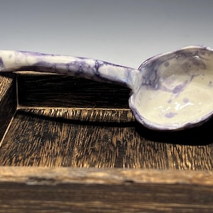 May include: A handmade ceramic ladle with a marbled purple and white design. The ladle rests on a dark wooden tray. The bowl of the ladle is off-white with purple accents.