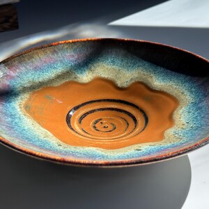 May include: A handmade ceramic serving bowl with a unique design. The bowl features a vibrant color palette, with a warm orange center and a gradient of blue, purple, and brown around the edges. The center has a spiral design.