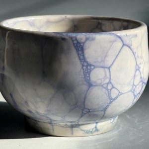 May include: A small ceramic bowl with a rounded shape and a smooth, glossy finish. The surface features a white base color decorated with a blue bubble-like pattern that creates a network of interconnected circles across the exterior and interior.