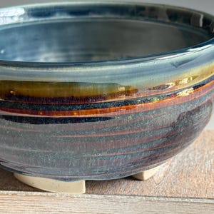 May include: A handmade ceramic bowl with a blue, brown, and orange glazed finish. The bowl has a rounded shape with a wide opening and sits on three small, off-white feet. The glaze creates a textured, layered effect, adding depth and visual interest.
