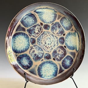 May include: A ceramic plate with a unique design. The plate features a pattern of blue and white circular shapes against a tan background, with a dark brown rim. The plate is displayed on a black metal stand.