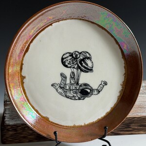 May include: A round ceramic plate with a cream-colored center and a wide, iridescent copper-toned rim. The center features a black line-art illustration of an astronaut floating while holding a cluster of balloons shaped like various planets.