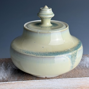 May include: A ceramic jar with a lid. The jar is a pale yellow color with a glossy finish. A band of turquoise-colored speckles encircles the jar. The lid has a small, decorative finial. The jar is sitting on a wooden surface.