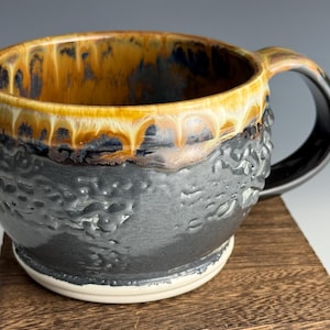 May include: Handmade ceramic mug with a textured, dark gray exterior and a glossy, multi-toned interior featuring shades of yellow, brown, and blue. The mug has a black handle and a white base, perfect for coffee or tea.