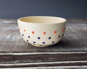 May include: A small, white ceramic bowl with a speckled pattern of blue and orange dots.