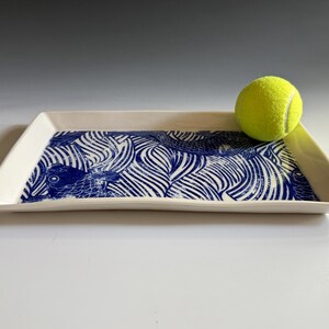 Pu&ograve; includere: Un vassoio rettangolare in ceramica con base bianca e una stampa grafica blu raffigurante pesci stilizzati e motivi a onde. Una pallina da tennis giallo brillante poggia sul lato destro del vassoio, fornendo una scala visiva delle dimensioni.